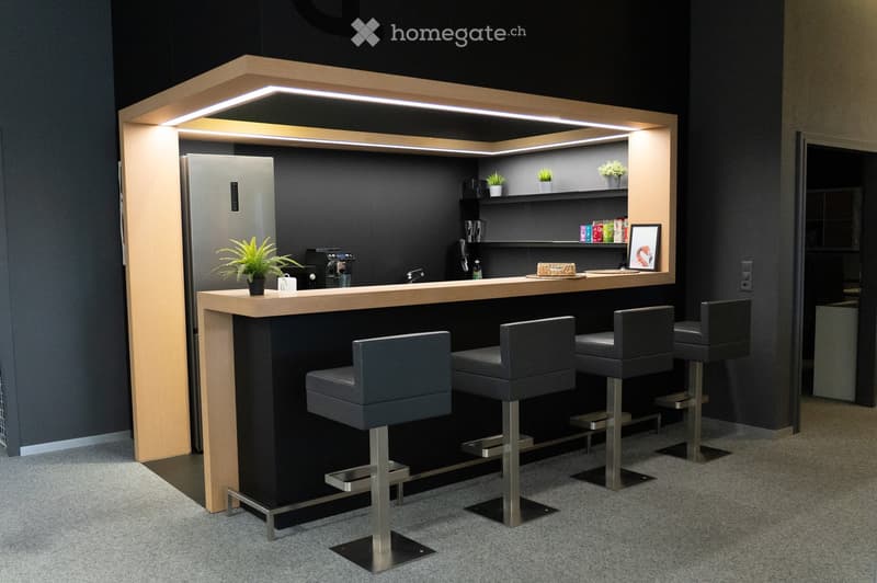 Modern bar area with a sleek wooden counter, four grey bar stools, a refrigerator, coffee maker, small potted plants, and shelves holding bottles and a picture frame.