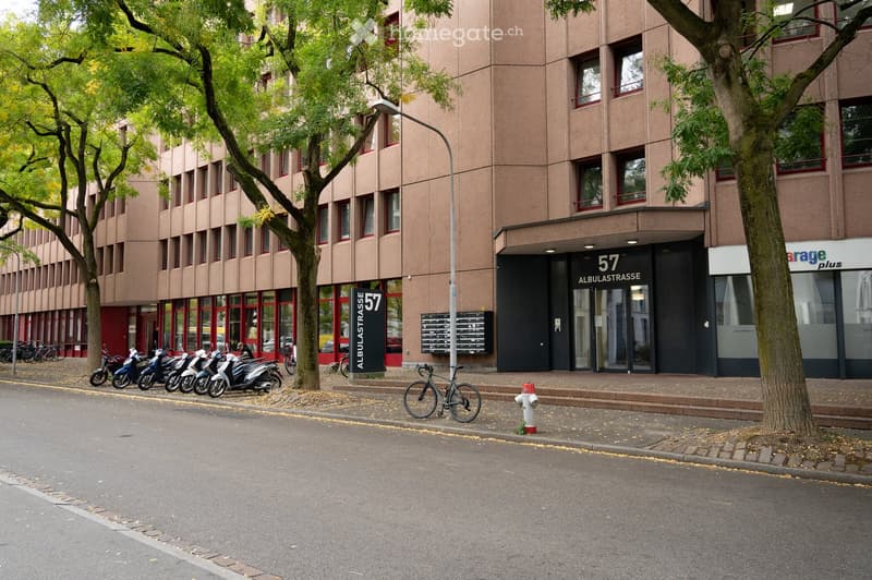 Multi-story building with red-framed windows and a modern entrance labeled '57 Albulastrasse'. Several scooters and bicycles are parked along the sidewalk, with trees lining the street.
