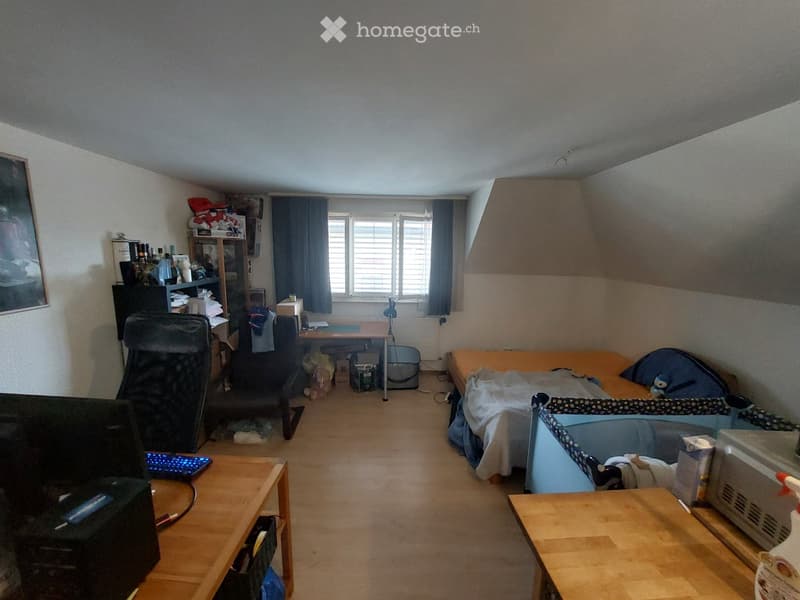 A small, furnished bedroom with a bed, desk, chairs, and a baby cot. The room features wooden flooring, a window with curtains, and various personal items on shelves and tables.