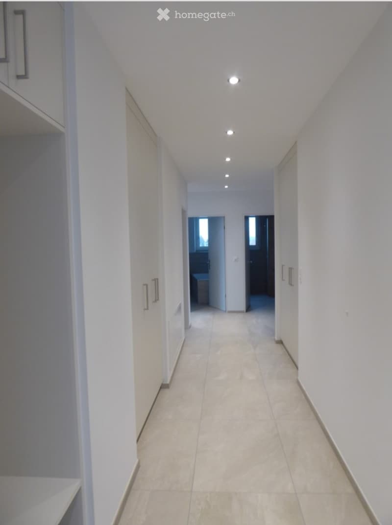 Empty hallway with white walls, tiled floor, and recessed ceiling lights. Doors lead to other rooms at the end.