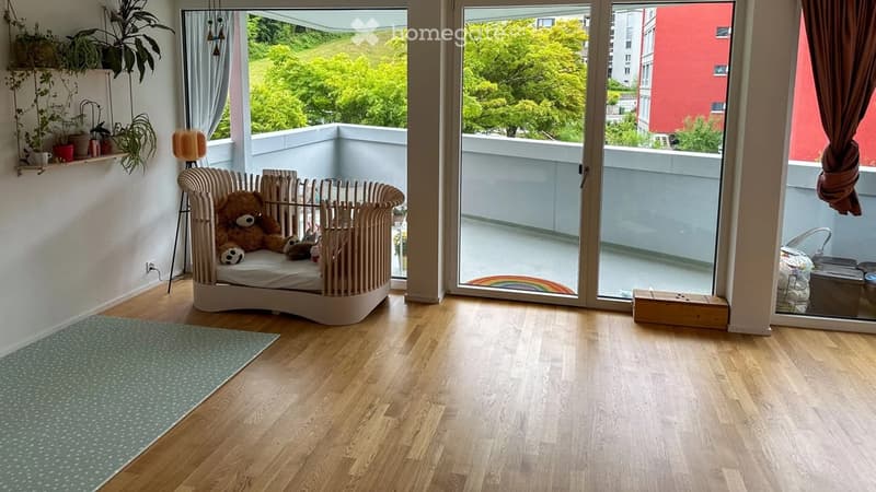 Bright, airy room with wooden flooring, large windows, and a balcony. Features a crib, a teddy bear toy, a green rug, and a shelf with plants and a lamp.