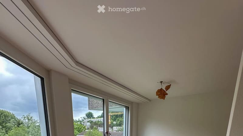 Empty room with large windows offering a view of the city and greenery, featuring a modern ceiling design and a decorative leaf-shaped light fixture.