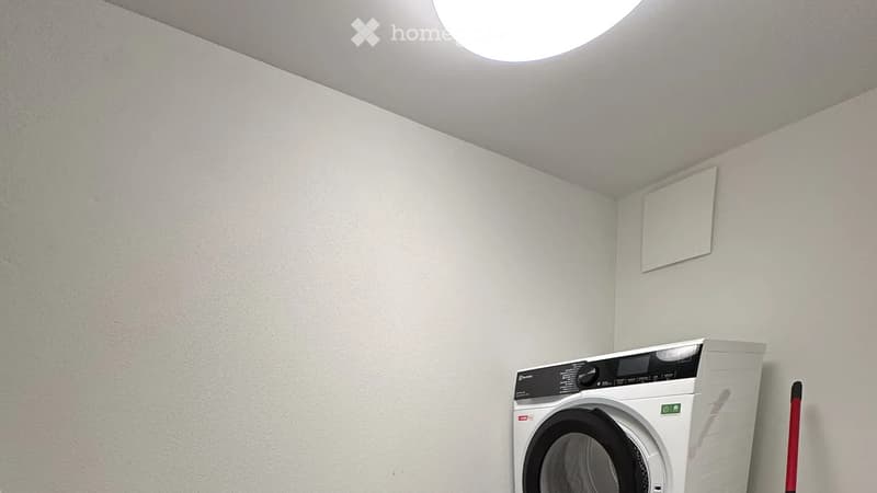 A small, clean room with a modern washing machine, white walls, and a skylight window on the ceiling.