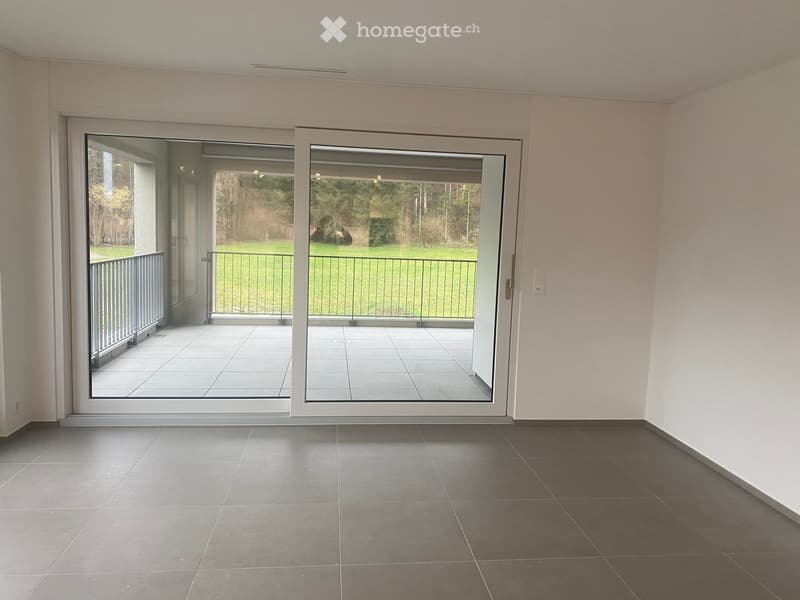 Empty room with grey tiled flooring, white walls, and large sliding glass doors leading to a balcony with a metal railing and a view of a green outdoor area.