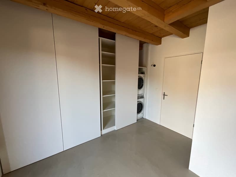 Empty room with white walls, wooden ceiling beams, and a built-in closet with shelves. A washing machine is visible in the corner.