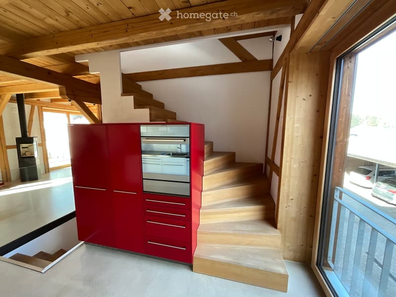 Modern kitchen with red cabinetry, built-in oven, sink, and staircase leading to an upper level. Exposed wooden beams and a fireplace are visible.