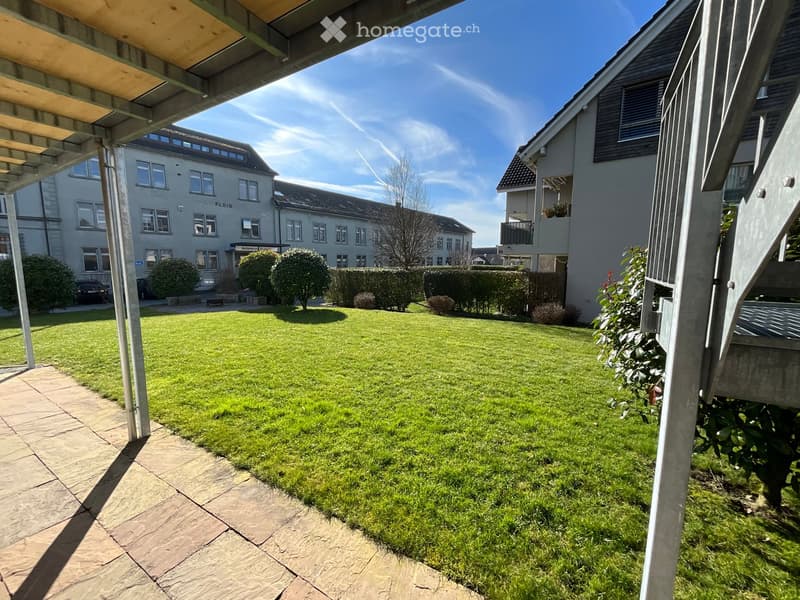 Well-maintained lawn with trimmed hedges and small trees, surrounded by residential buildings and a covered patio area.