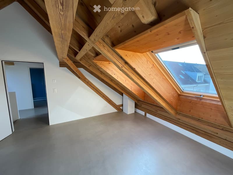 An empty attic space with exposed wooden beams, a skylight window, and a concrete floor.