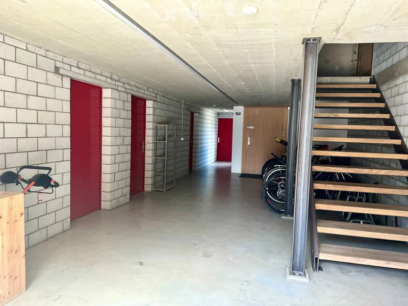A modern, clean corridor with concrete floors and walls, featuring a wooden staircase on the right and several red doors along the left side. Bicycles are parked near the staircase, and a small metal shelf is visible against the wall.
