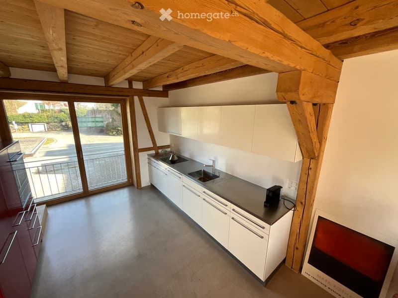 Modern kitchen with white cabinets, stainless steel sink, and dark countertop. Exposed wooden beams on the ceiling and large windows provide natural light.