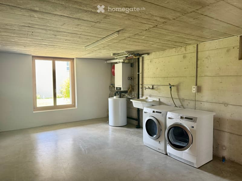 Utility room with concrete walls, wooden ceiling, and large window. Contains a washing machine, dryer, water tank, and sink.