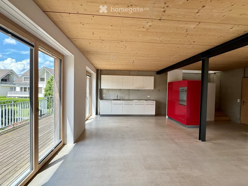 Modern, open-plan kitchen area with white and red cabinets, a sink, and an oven, connected to a spacious living area with wooden ceilings and large windows overlooking a balcony.