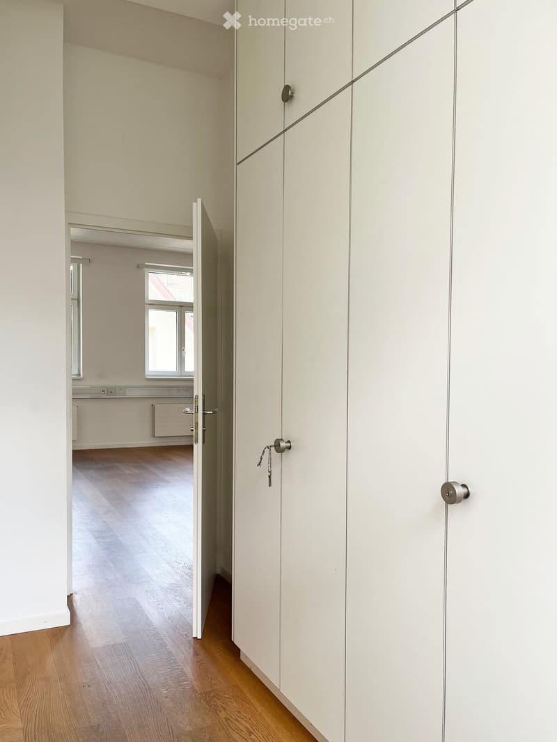 Empty hallway with light-colored wooden flooring and white walls, featuring an open door leading to another room with a window and radiator.