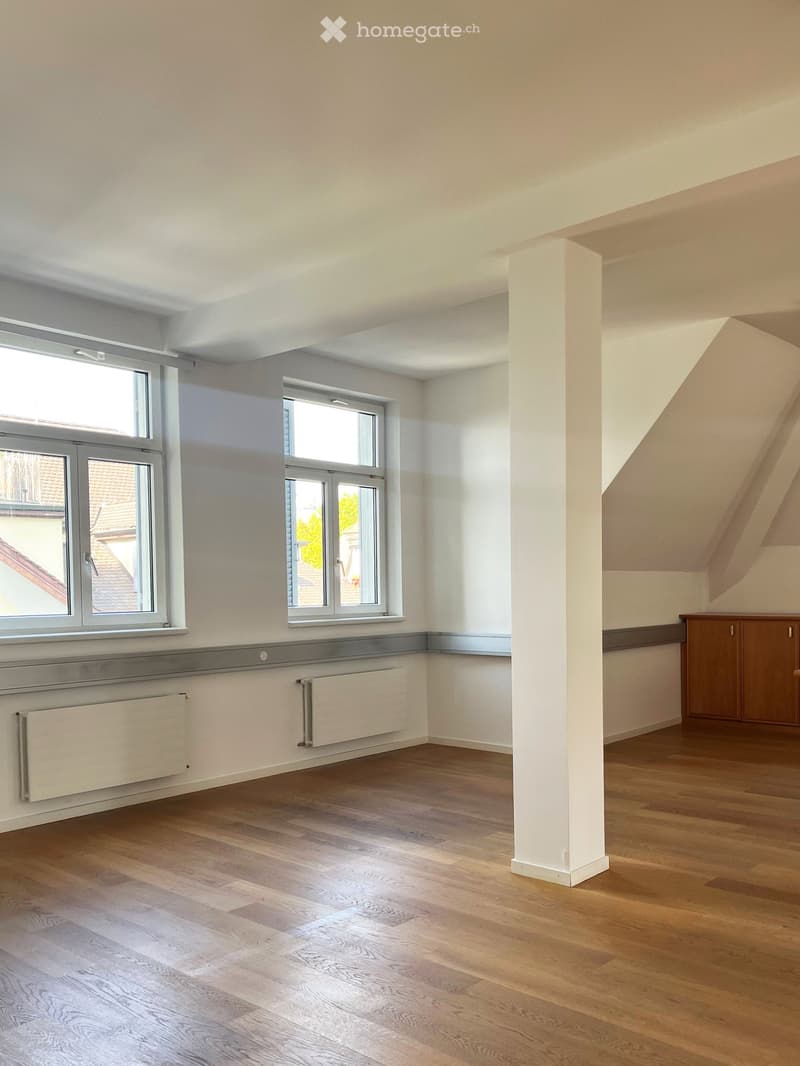 Empty room with wooden flooring, white walls, two windows, radiators, and a small wooden cabinet in the corner.