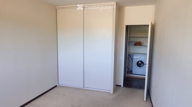 Empty room with beige walls, carpeted floor, and a large closet with sliding doors. An adjacent laundry area contains a washing machine and folded towels.