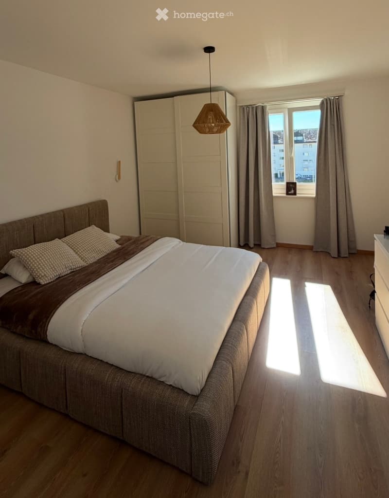 A modern bedroom with a large bed, pillows, a blanket, a wardrobe, and a hanging lamp. Natural light streams in from a window with curtains.