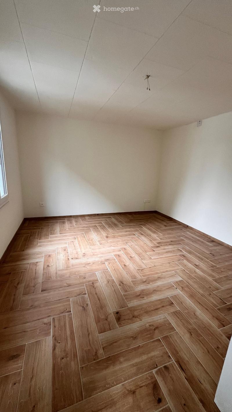 Empty room with light wooden herringbone flooring and white walls, featuring a small window on one side.