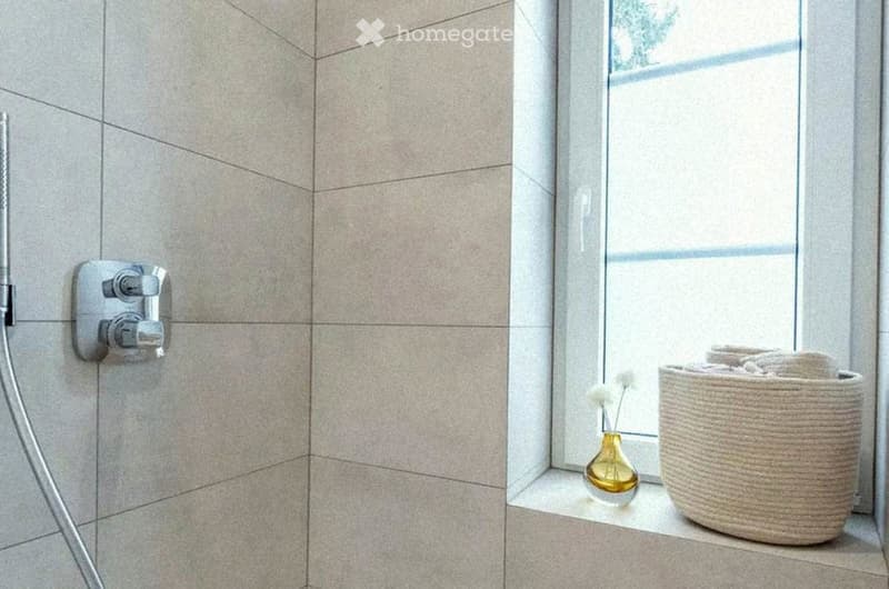 Modern bathroom with beige tiled walls, a showerhead and faucet on the left, and a window sill with a decorative vase and woven basket on the right.