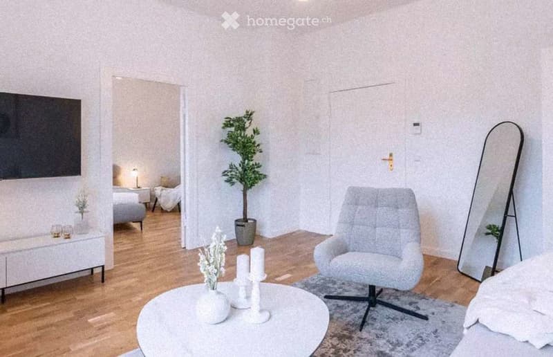 Modern living room with a grey sofa, coffee table, armchair, large mirror, table lamp, decorative plant, and candles. The room features light wooden flooring and white walls.