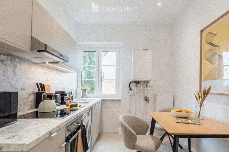 Modern kitchen with white cabinets, marble countertops, and stainless steel appliances. Features a breakfast area with a wooden table, two chairs, and a bowl of fruit on the table.