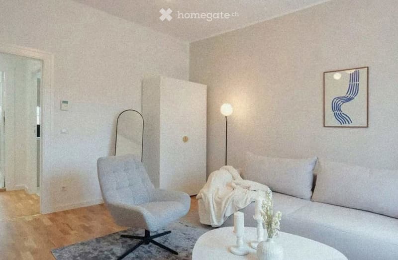 Modern living room with light-colored walls, wooden flooring, a grey sofa with a blanket, a matching armchair, a white coffee table with candles, and a floor lamp.