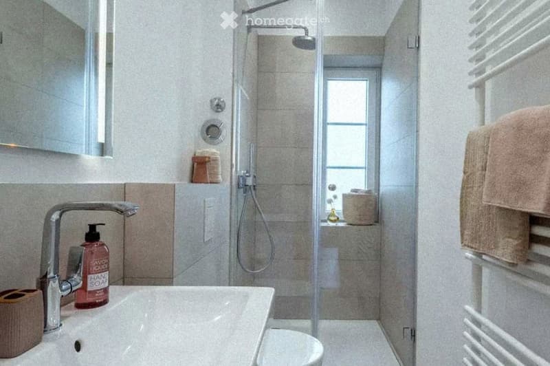 Modern bathroom with a white sink, chrome faucet, hand soap bottle, toilet, glass-enclosed shower, and a towel on a rack.