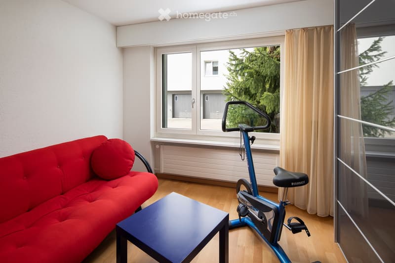 A modern living room with a red sofa, a black coffee table, and a blue exercise bike positioned near a large window with beige curtains.