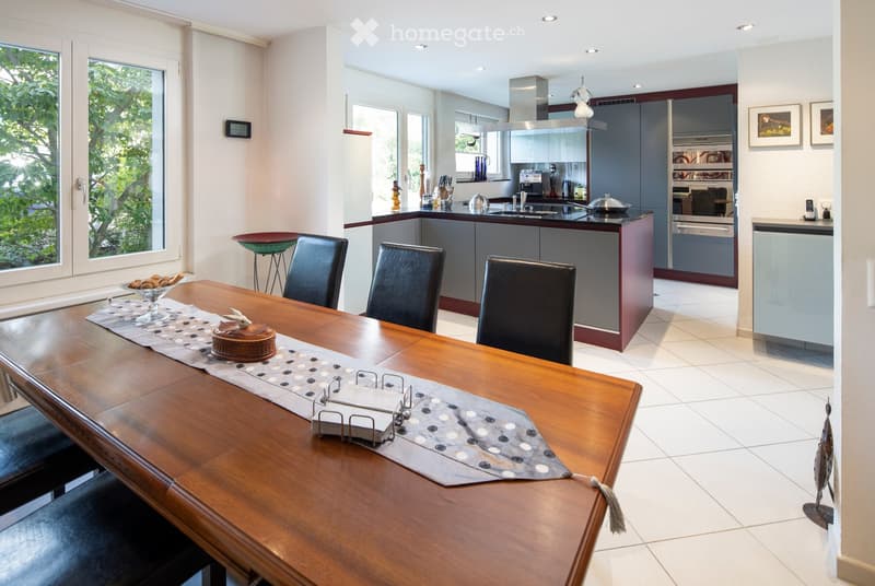 Open-plan dining and kitchen area with a wooden dining table, four black chairs, a modern kitchen with grey cabinets, a stainless steel range hood, and various kitchen appliances.