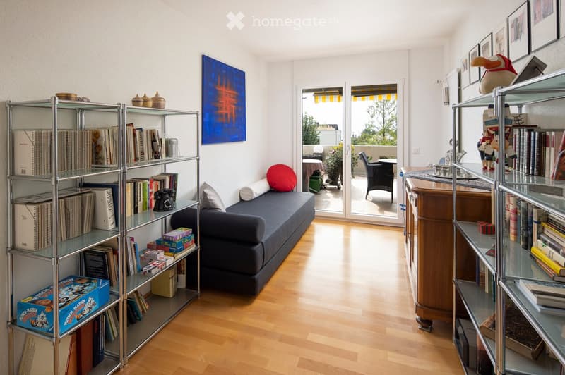 Modern living room with wooden floor, large window opening to a balcony, sofa, shelves filled with books and decorative items, and a small table with a laptop.