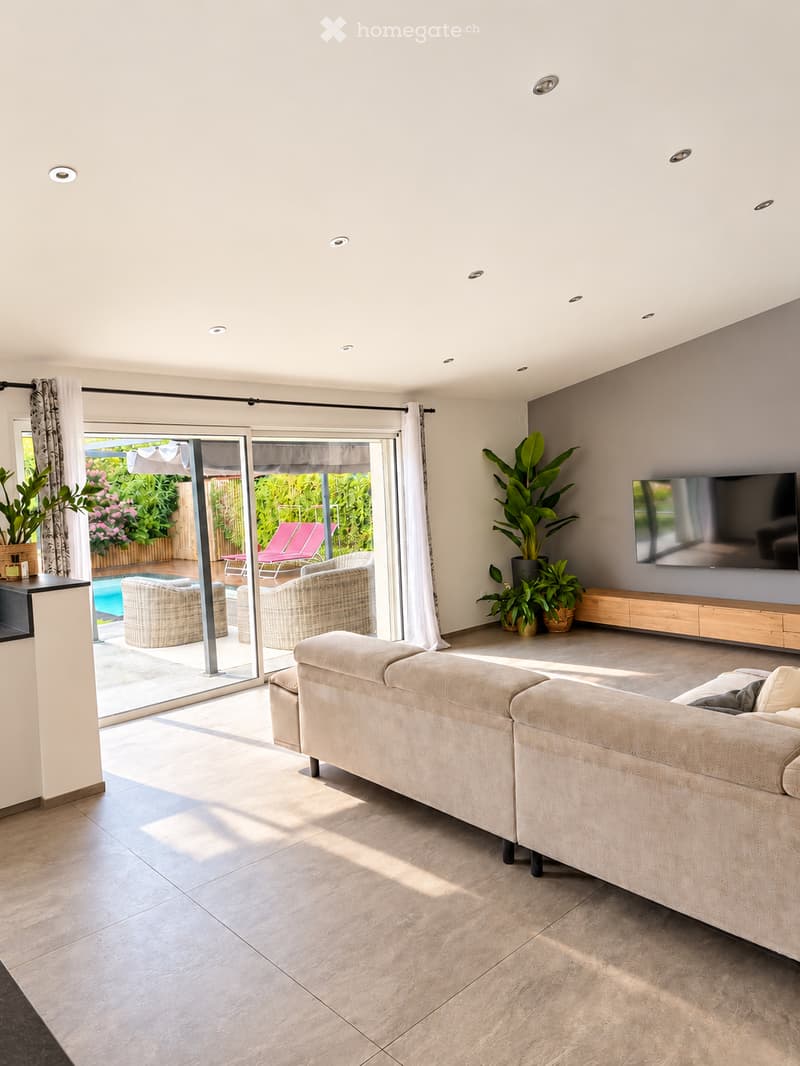 Modern living room with large sliding glass doors leading to a patio, a beige sectional sofa, a flat-screen TV mounted on the wall, and several potted plants.