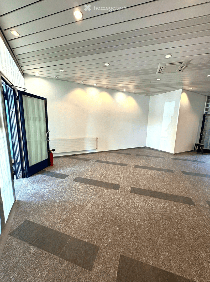 Empty room with polished tile floor, white walls, and recessed lighting. Features a glass door with a blue frame and a red fire extinguisher near the entrance.