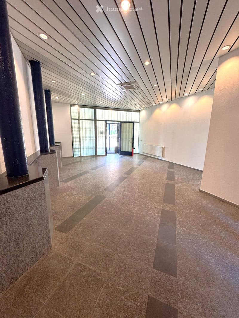 Modern hallway with polished tile flooring, white walls, and recessed lighting. Features a large glass door at the end leading outside, with a radiator on the right wall and a dark pillar on the left.