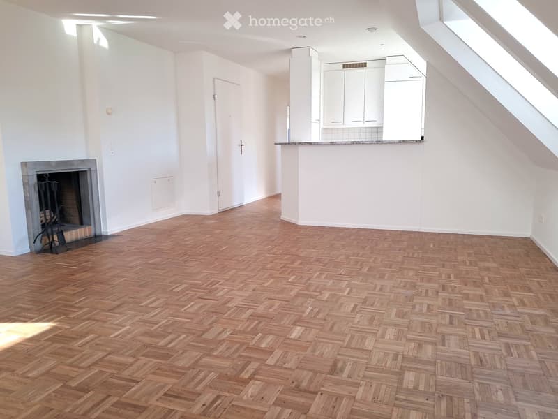 Empty room with wooden herringbone flooring, white walls, fireplace, and a small kitchen area with cabinets and a countertop.