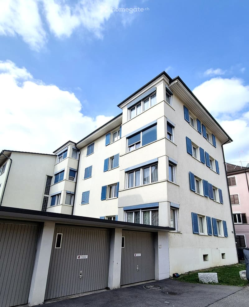 Four-story residential building with light beige facade, blue shutters, and multiple windows. Features a covered parking area with two garage doors.