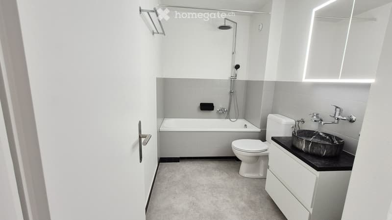 Modern bathroom with a bathtub, toilet, sink, and showerhead. Features a large mirror, towel rack, and sleek fixtures.