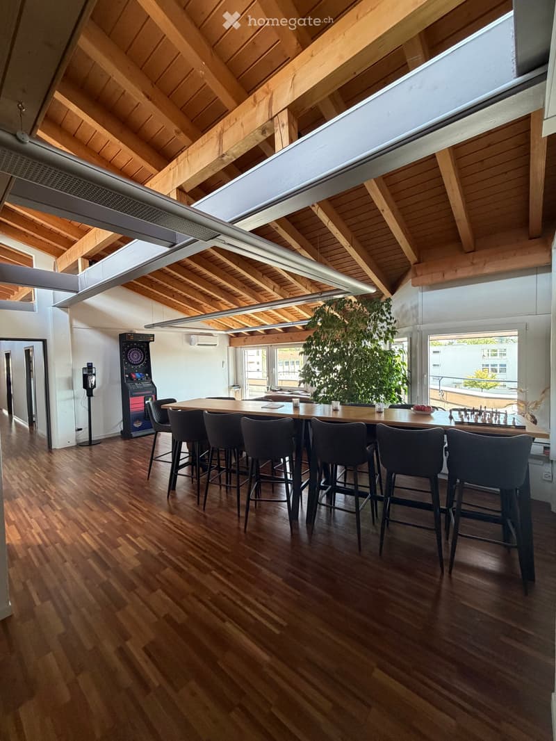 A spacious, modern room with wooden flooring and exposed wooden beams on the ceiling. A large central table is surrounded by several high-backed chairs. An arcade machine is positioned against the wall, and a chessboard is set up on the table. A large gre