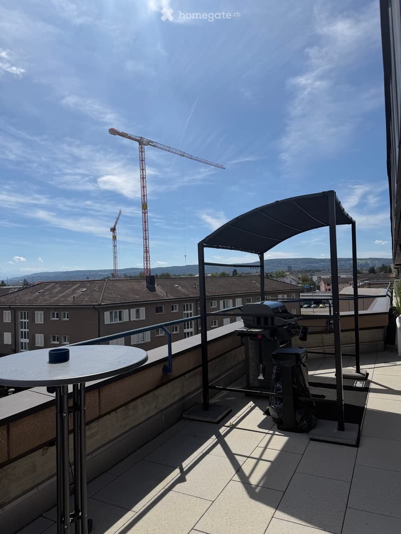 Balcony with a round table, umbrella, and grill, overlooking a cityscape with cranes and buildings.