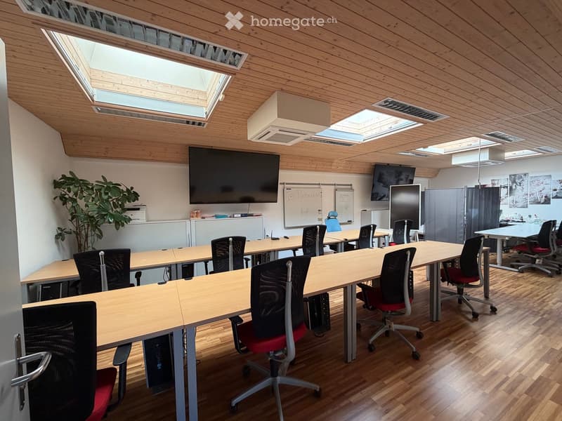 Bright, modern office space with wooden floors, large skylights, and multiple tables with chairs arranged for meetings. Features include a large screen, a potted plant, and whiteboards.