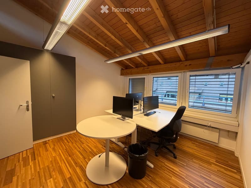 Modern office space with wooden flooring, white desk, round table, office chair, multiple computer monitors, and a trash can.