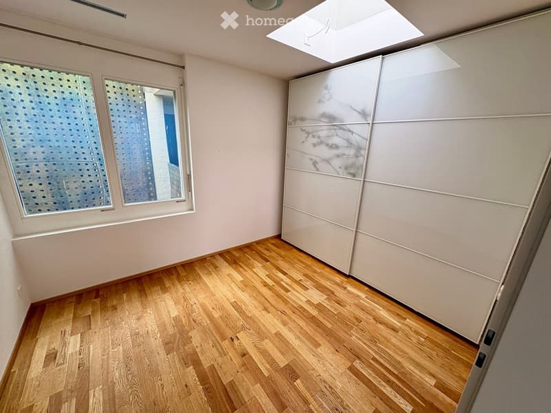 Empty room with light wooden flooring, white walls, and large windows with perforated glass panels. A white partition wall is visible on the right side.