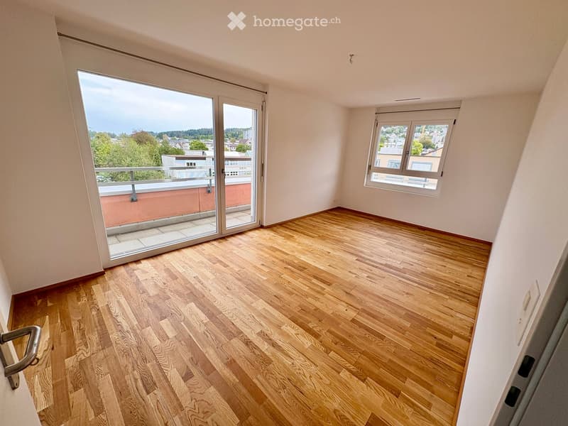 Empty room with wooden flooring, large sliding glass door leading to a balcony, and a window with a view of the city.