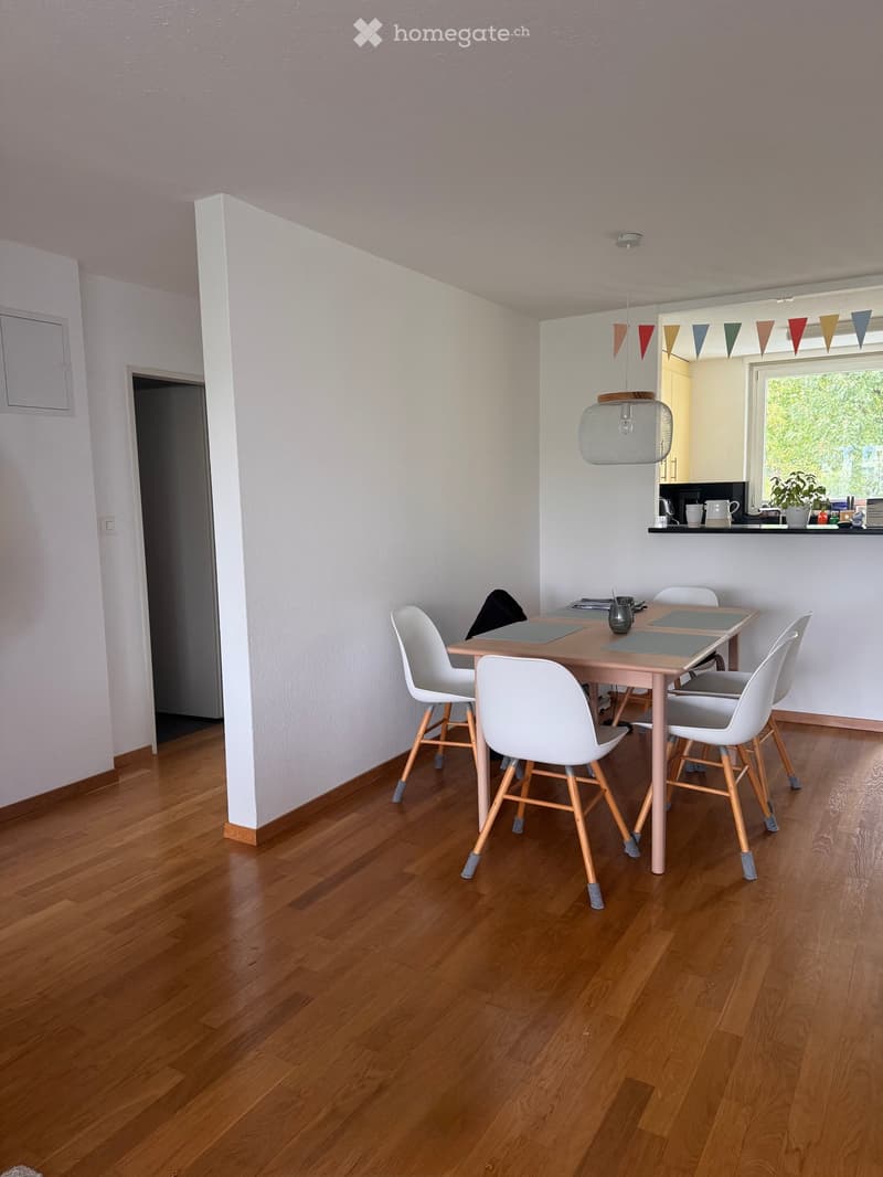 Dining area with a wooden dining table, white chairs, pendant light, and a view of a kitchen area with a window and countertop.