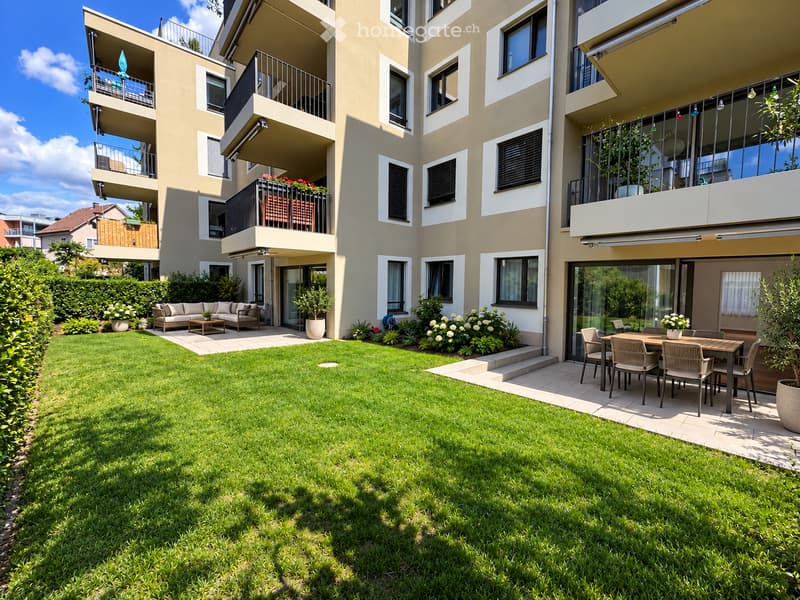 Modern residential building with multiple balconies, green lawn, patio furniture, and outdoor dining area.