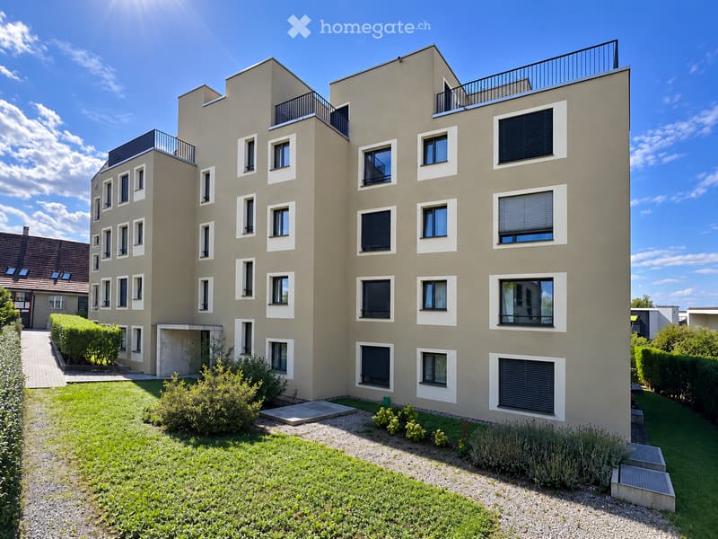 Modern multi-story residential building with beige facade, multiple windows, balconies, and surrounding greenery.