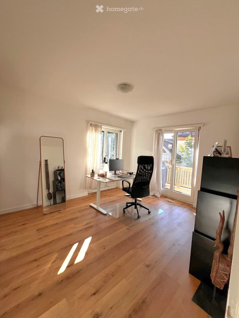 A well-lit room with wooden flooring, a large mirror, a desk with a chair, a TV, and a shelf with books and decorative items.