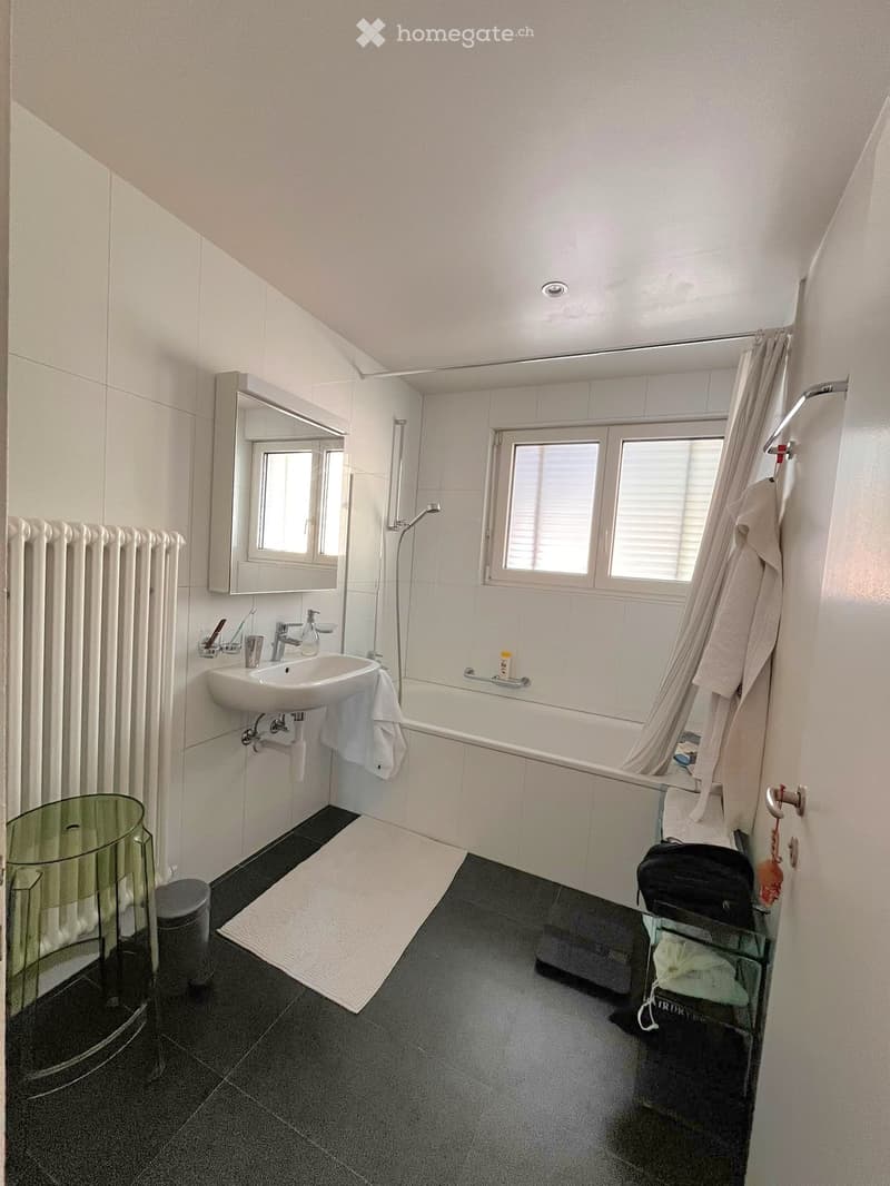 Modern bathroom with white tiles, a sink, bathtub, mirror, and a window. Includes a towel, cup, toothbrush, toothpaste, and a shower curtain.