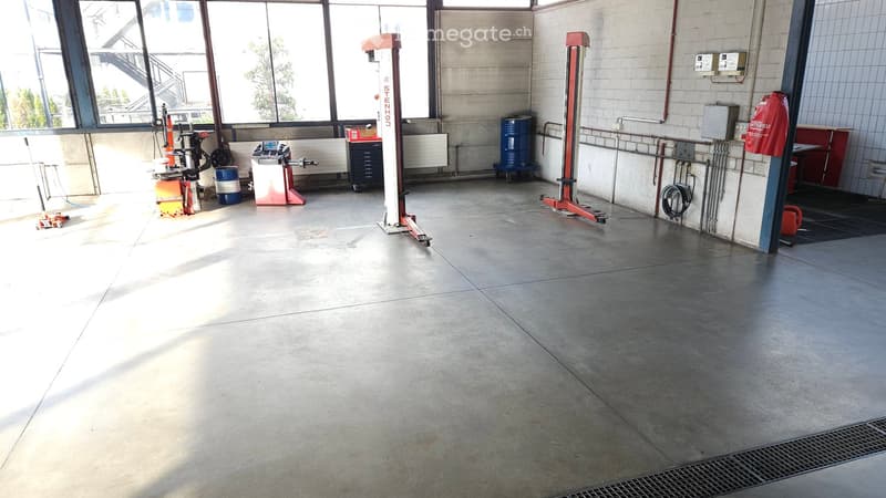 Empty garage with concrete floor, large windows, and various equipment including a hydraulic lift, tool cabinets, and storage barrels.