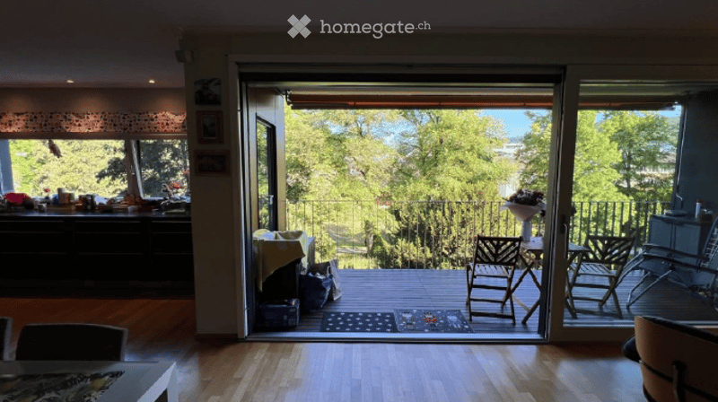 A modern, well-lit room with large windows and a sliding glass door leading to a balcony. The room features wooden flooring, a small table with chairs, and a cabinet with various items on top.