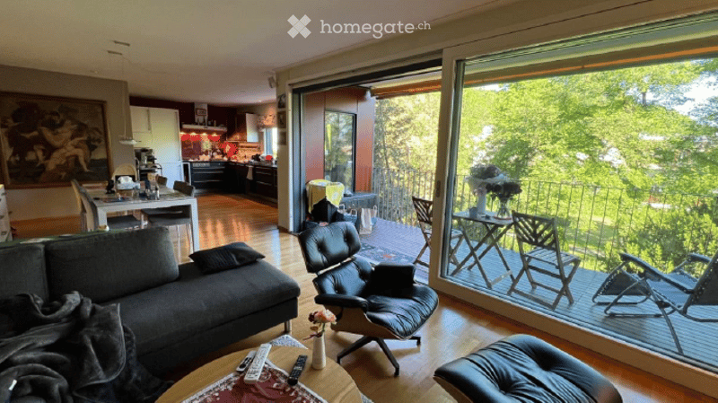 Modern living room with large windows and a balcony overlooking greenery. Features a grey sofa, coffee table with remote controls, leather chair, and a small table with a vase of flowers. The adjacent kitchen area is visible with a dining table and chairs