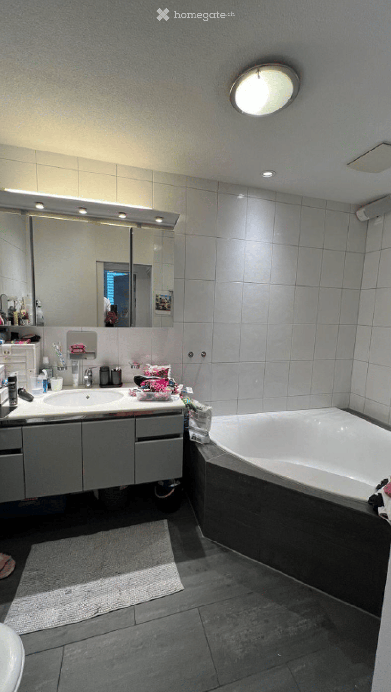 Modern bathroom with a large mirror, double sink, bathtub, and grey tiled walls and floor.